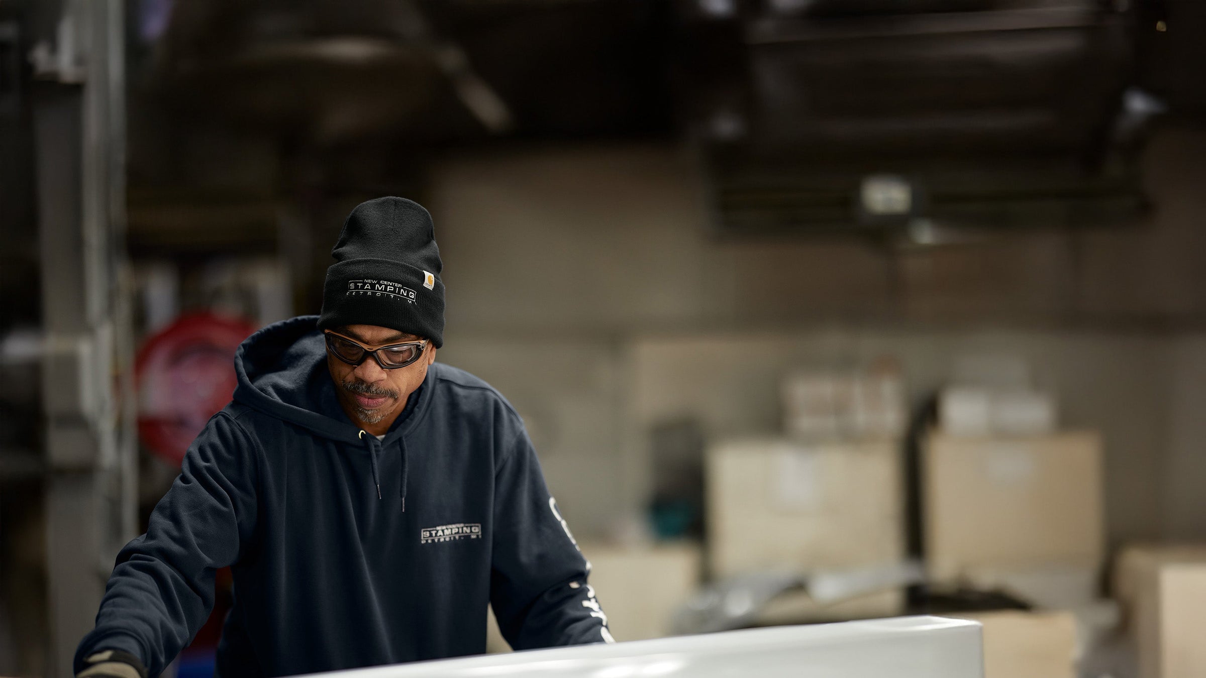 A worker wearing a black custom beanie and navy hoodie is focused on a task in an industrial setting. The background contains stacked boxes and equipment, indicating a workshop or warehouse environment.