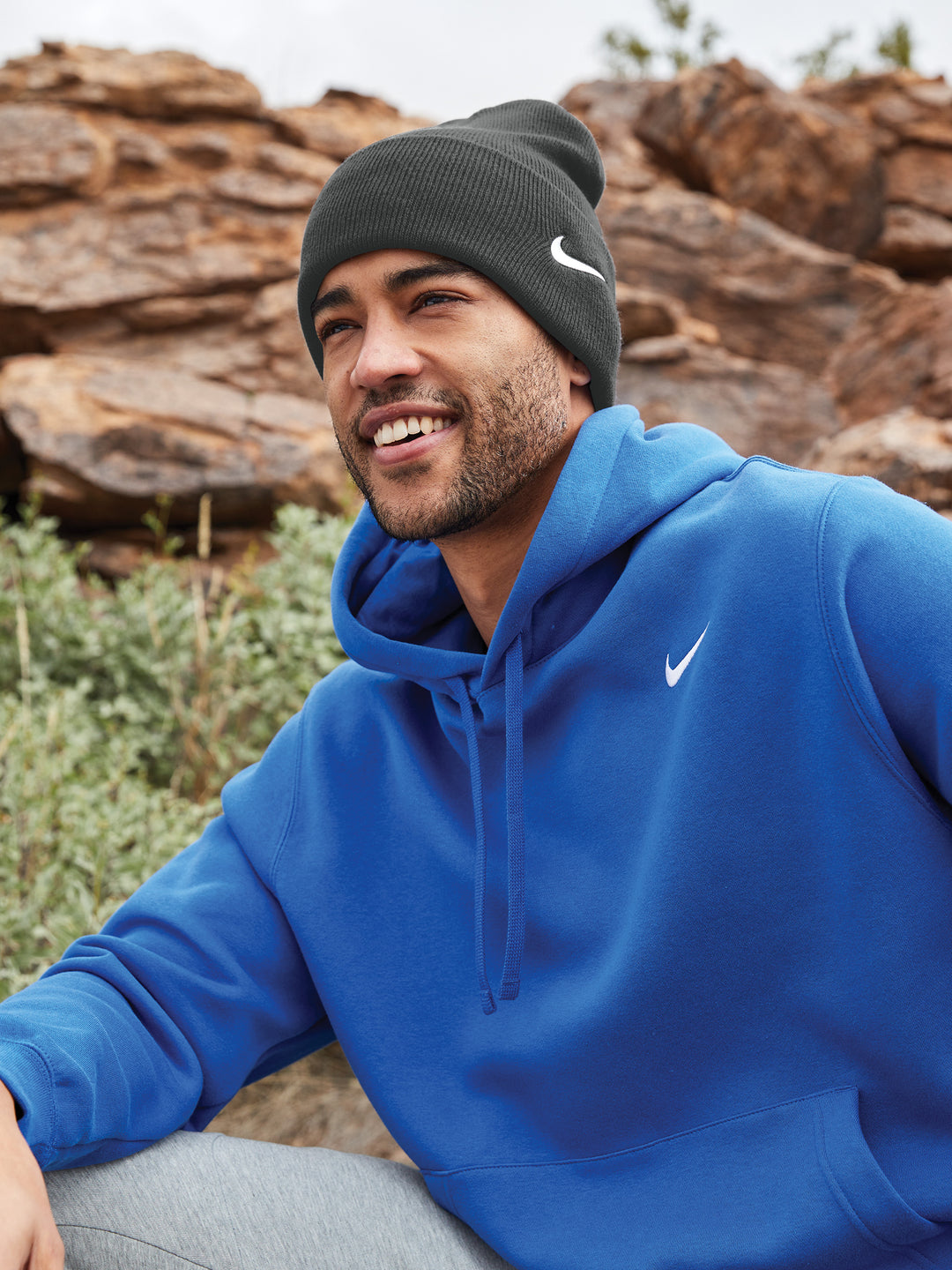 Man wearing a black Nike Team Cuffed Beanie with a white swoosh logo, paired with a blue hoodie. Outdoor setting with rocks and greenery.