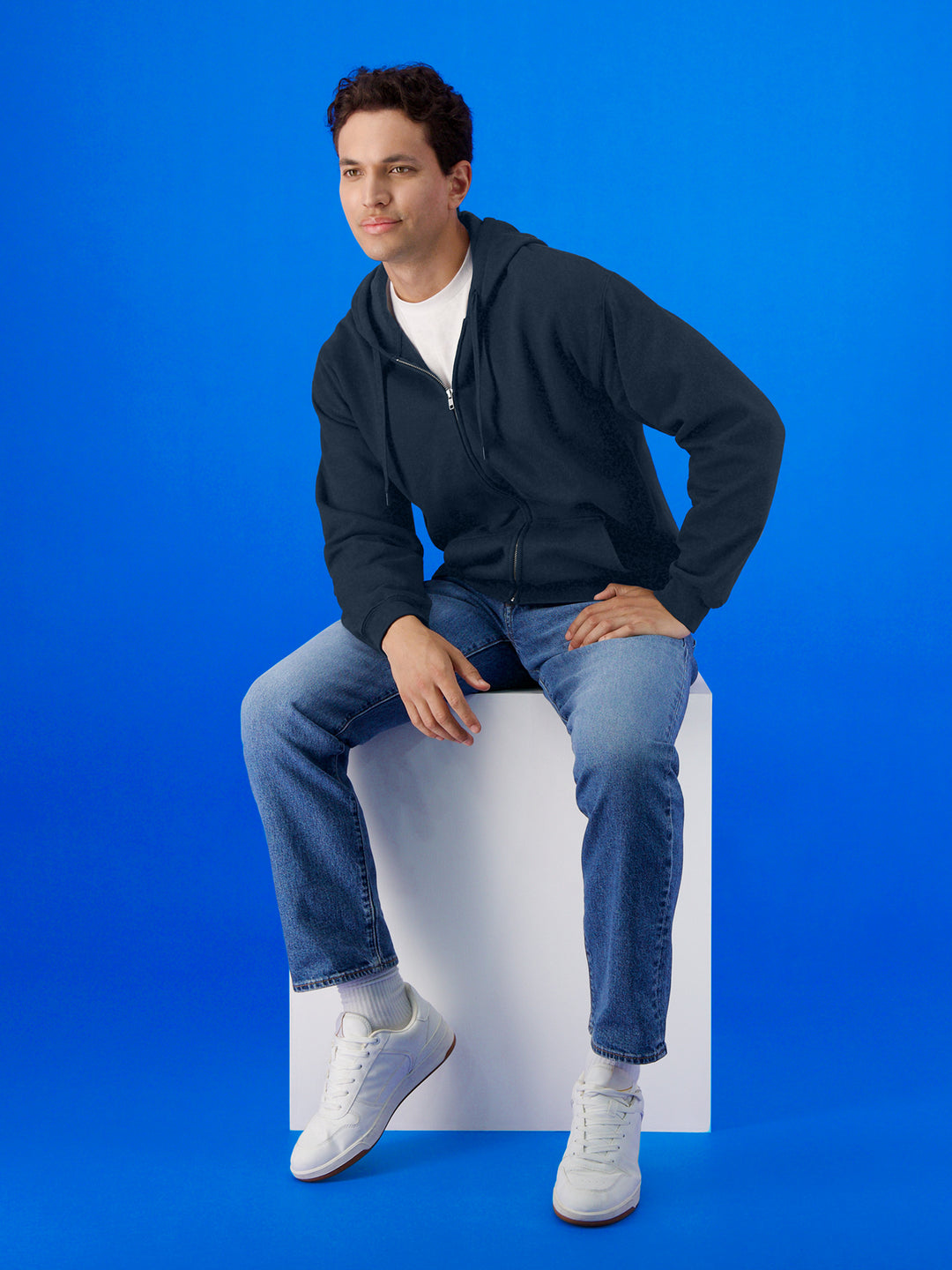 Model wearing a Gildan Softstyle Full Zip Hooded Sweatshirt in black, seated against a blue background. A versatile hoodie for layering and customization.