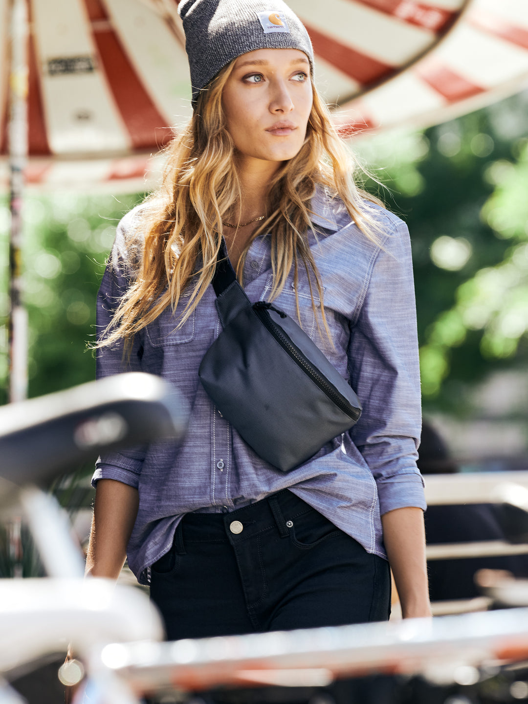 Woman wearing a Carhartt® Acrylic Knit Hat Beanie in gray, paired with a casual outfit and crossbody bag, outdoors under a striped umbrella.