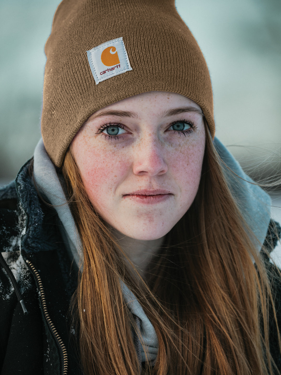 Woman wearing a Carhartt® Acrylic Knit Hat Beanie in brown, bundled up in winter clothing with snow on her jacket.