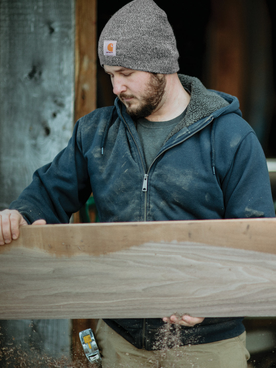 Man wearing a Carhartt® Acrylic Knit Hat Beanie in brown while working with wood, dressed in a rugged hoodie and workwear.